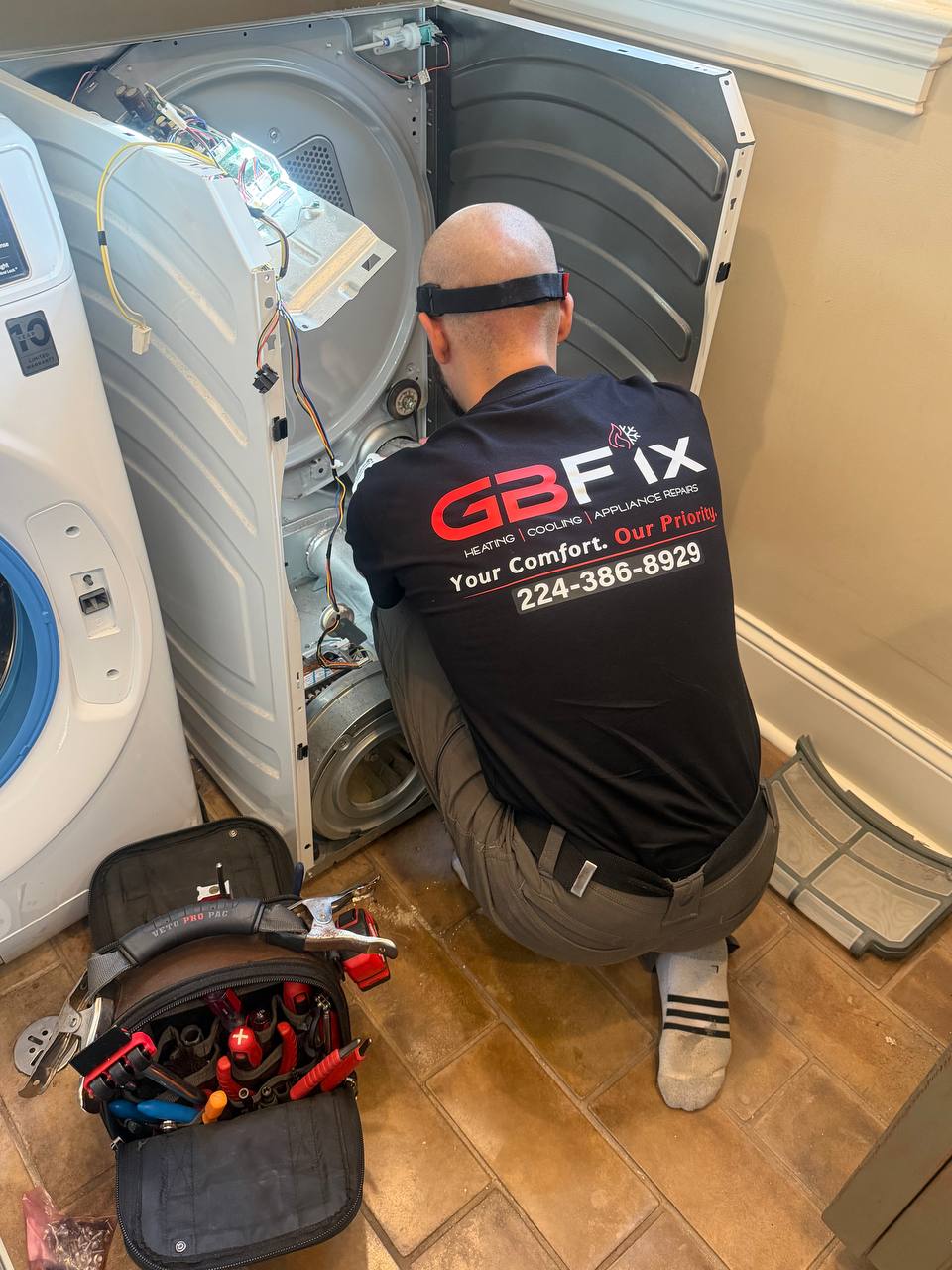 Technician kneeling while repairing a disassembled clothes dryer beside a washing machine